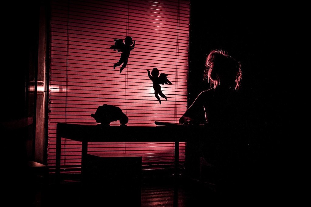 Σκοτεινή φωτογραφία όπου διακρίνεται η φιγούρα μίας κοπέλας που κάθεται δίπλα σε τραπέζι, ενώ στο φόντο οι φωτισμένες κόκκινες περσίδες φέρουν τις σκιές από δύο μικρούς αγγέλους. Η εικόνα συνοδεύει το κείμενο «Τόπος».