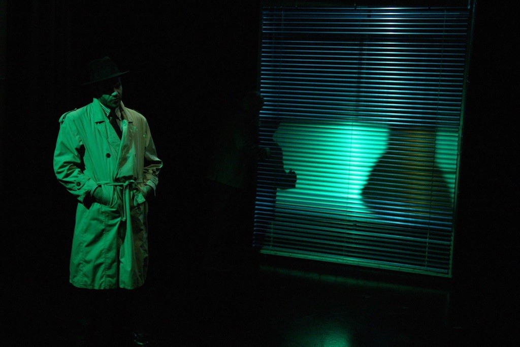 Ένας άντρας με καπαρντίνα και καπέλο στέκεται δίπλα σε κλειστές περσίδες